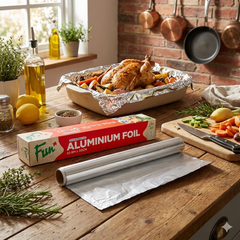 Aluminium foil roll on a wooden kitchen counter with a roasted chicken and vegetables.