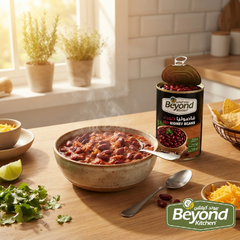 A bowl of chili with kidney beans sits on a wooden kitchen table beside an open can of Beyond Kidney Beans. The scene includes a spoon, lime, cilantro, shredded cheese, tortilla chips, and a bright kitchen background with potted plants and a window.